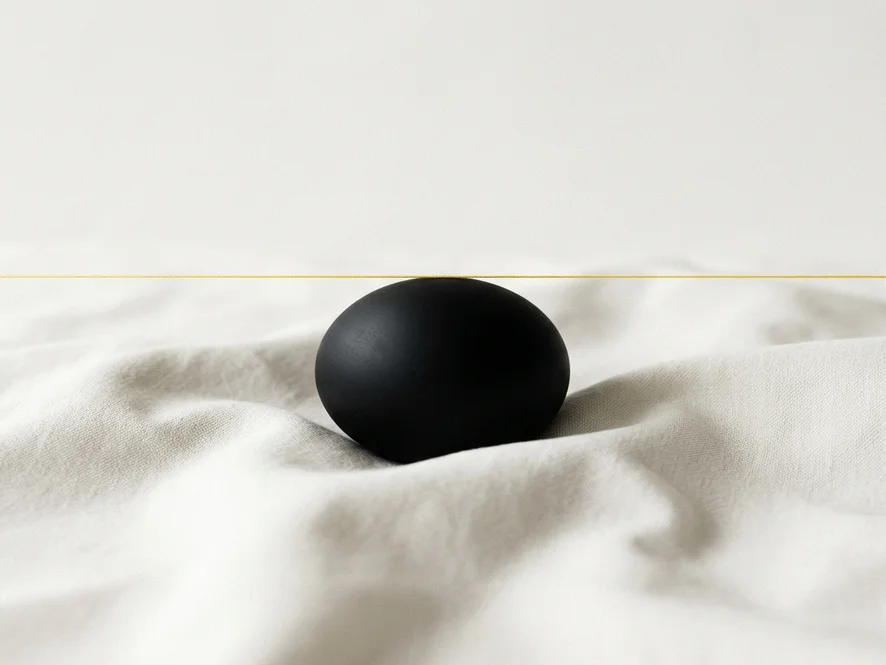 Swiss-editorial minimal still life showing a matte black sphere on softly folded bone-white fabric with a thin yellow thread representing ideas igniting.